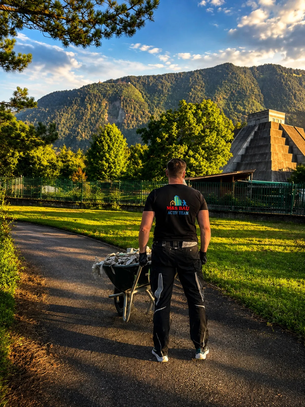 Person im M&S Bau Activ Team T-Shirt schiebt eine Schubkarre mit Bauschutt über einen Weg, im Hintergrund Berge im Abendlicht